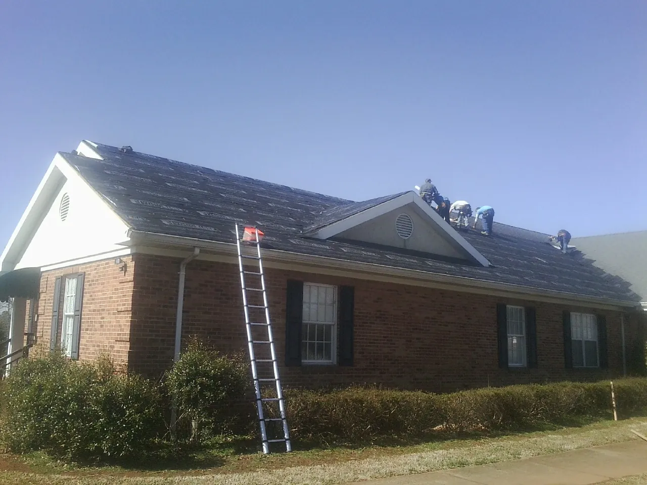 Crew tearing shingles off a brick ranch roof with a ladder set — Emergency Roof Repair work by our crew in Mc Cool Junction