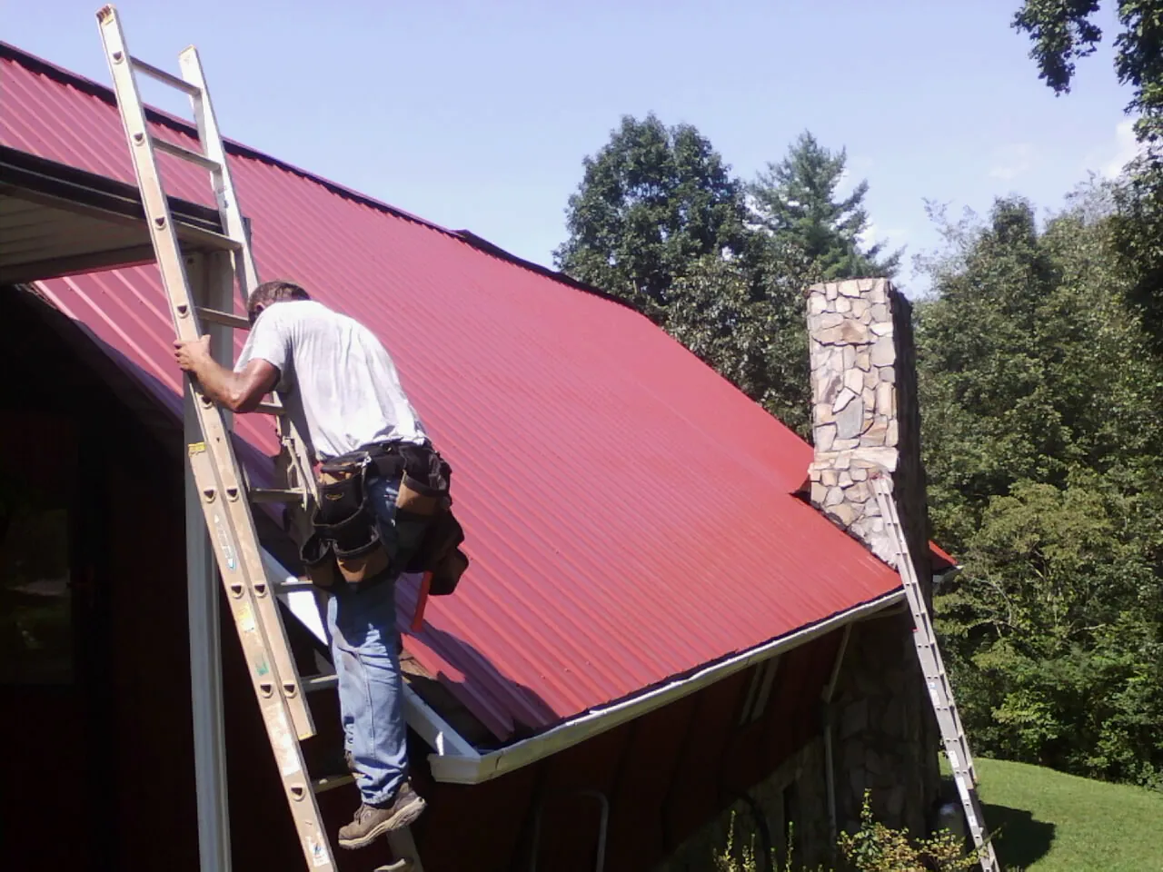 Roofer on a ladder beside a red metal roof and stone chimney — Soffit & Fascia Repair by Roofing Brothers in Mc Cool Junction